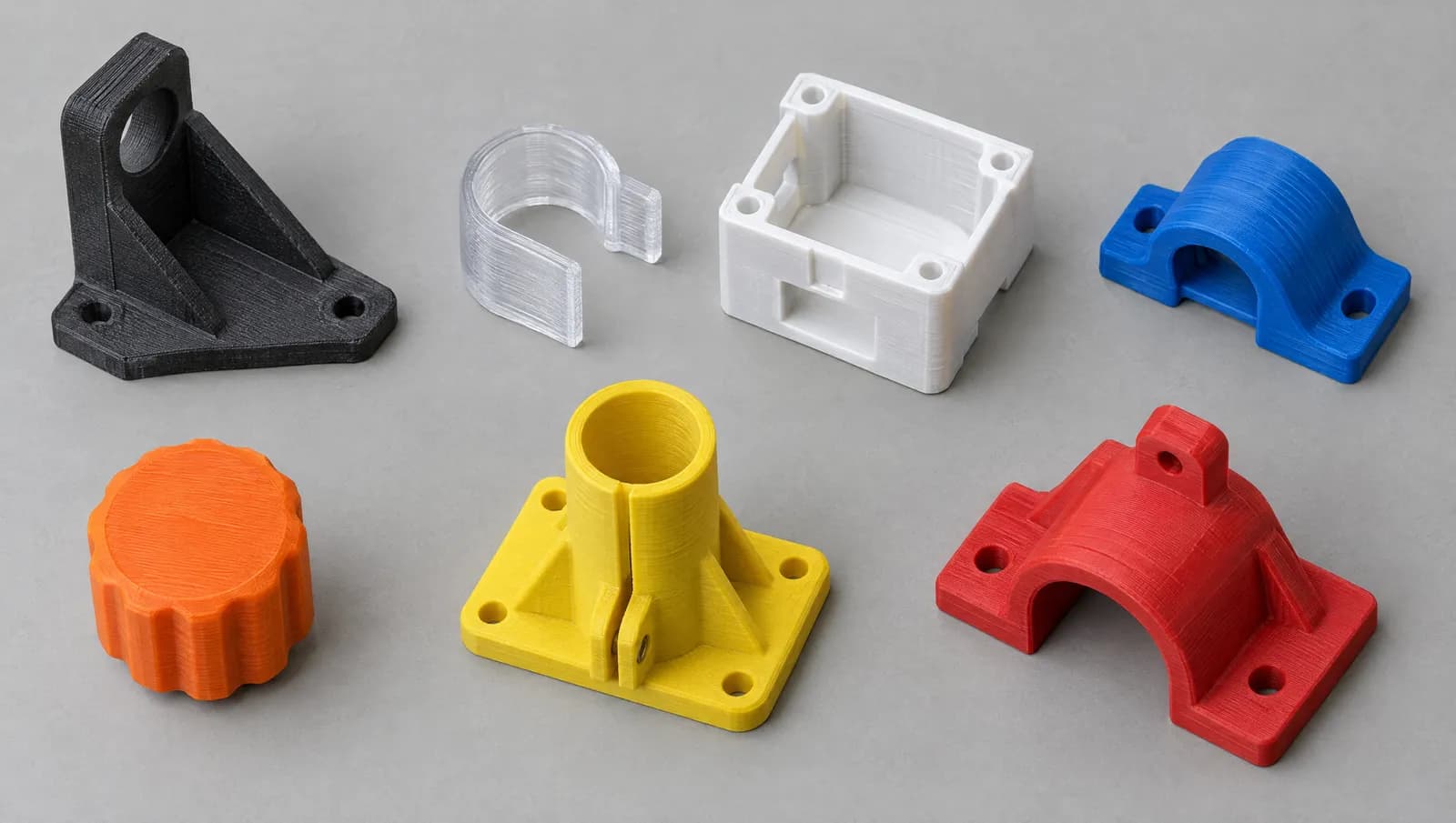 Seven printed parts on a grey surface: a black bracket, a clear clip, a white box, a blue clamp, an orange knob, a yellow mount and a red clamp.